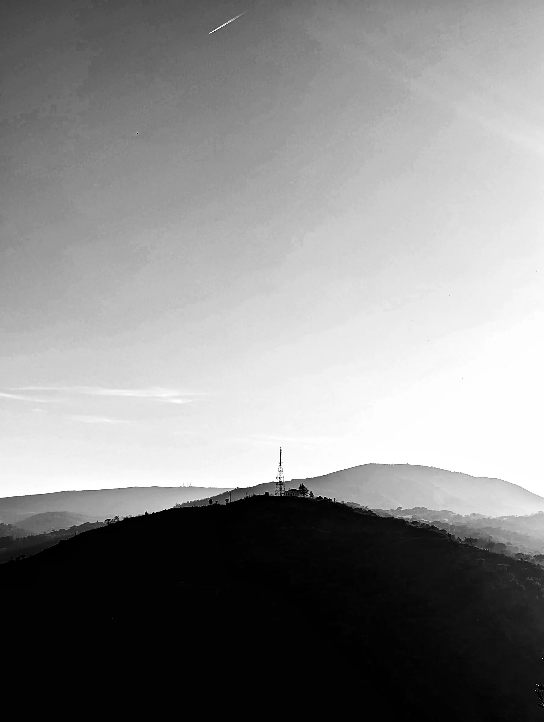 A black and white view of a dark wooded hill with a lattice radio mast on its summit, hazy ridges fading behind it and a plane's contrail cutting across the empty sky
